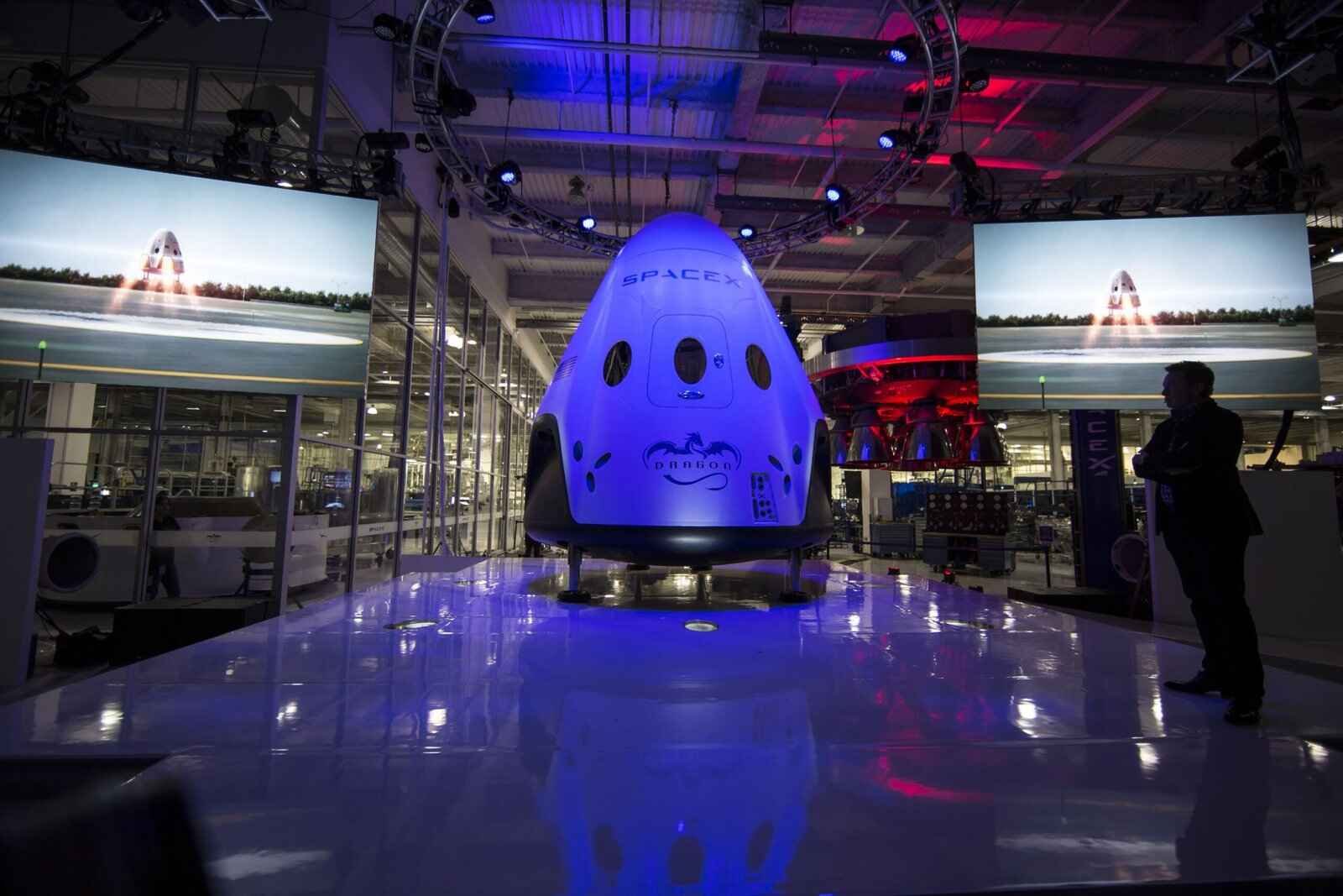 spacecraft-693221-scaled (1) SpaceX Dragon capsule on display with launch visuals and observer nearby.<br />